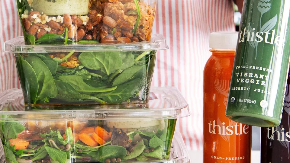 Stacked Thistle meal containers of salads and grain bowls beside Thistle cold-pressed juices